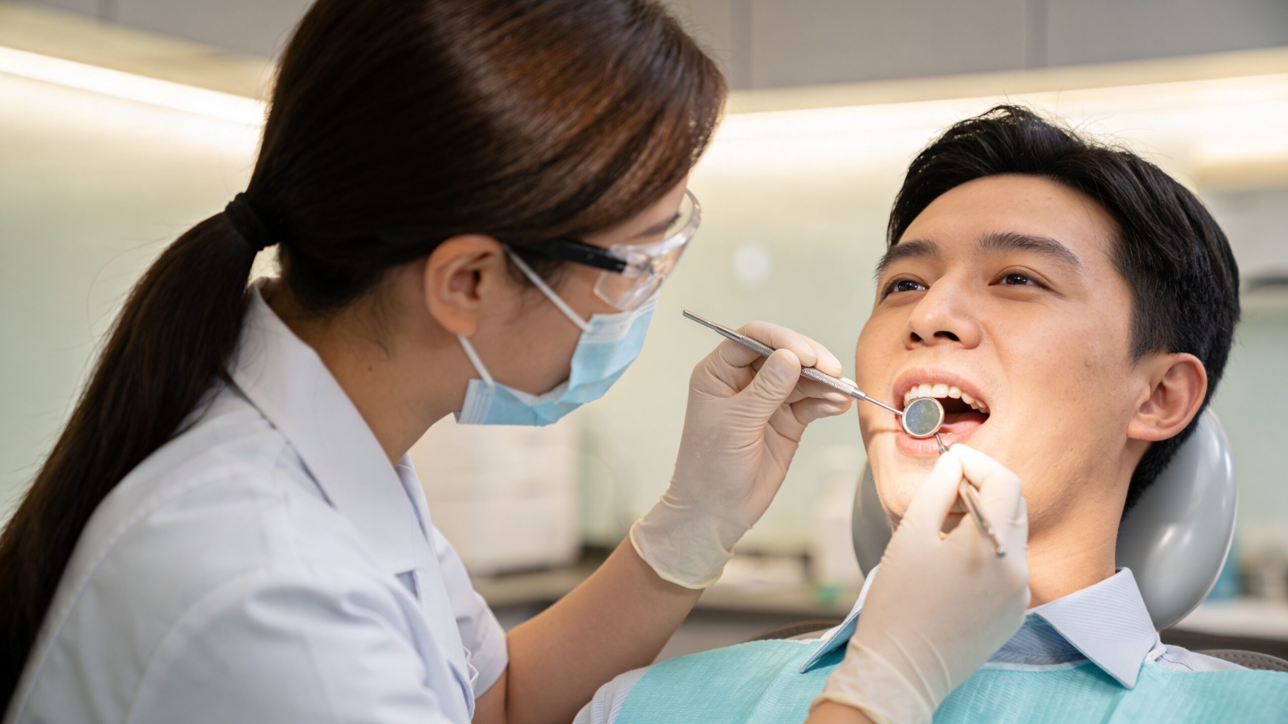 A female dentist in protective gear examines a male patient's teeth using a dental mirror and probe.